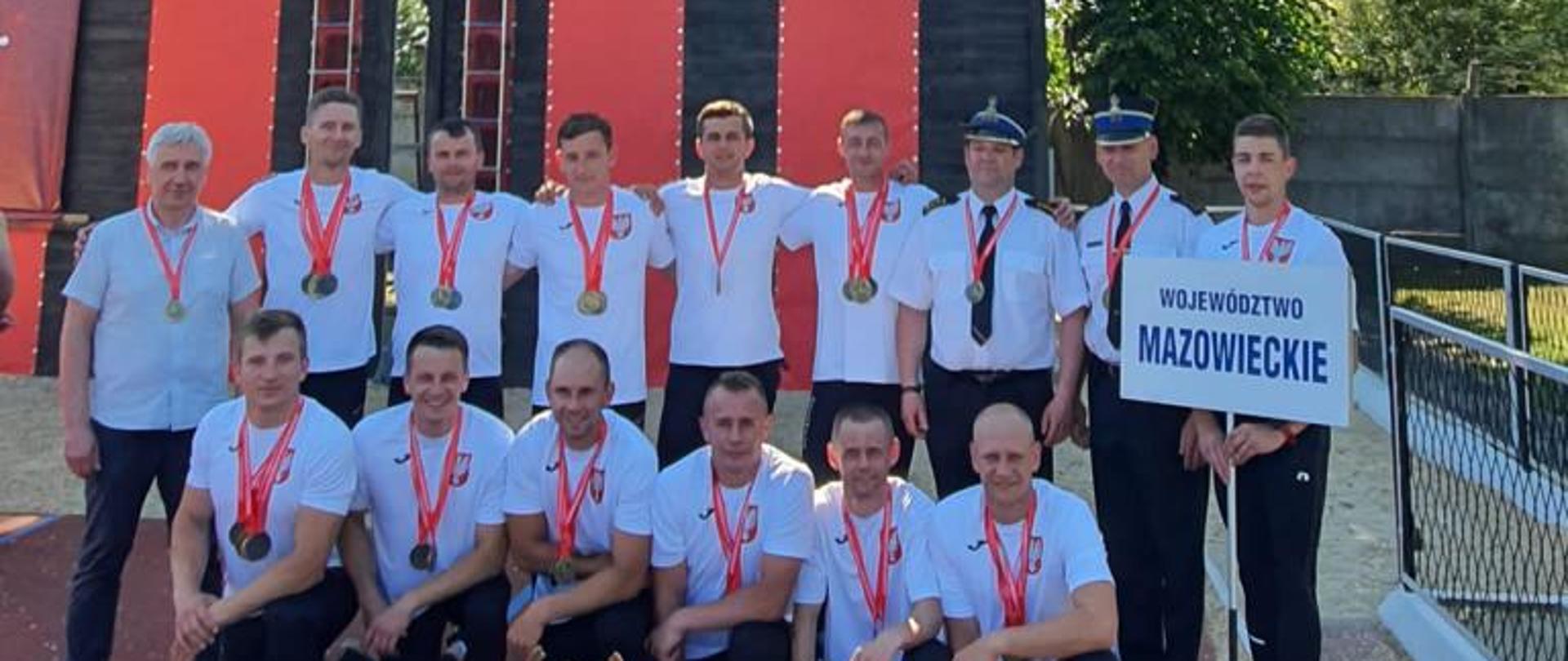 Mazowsze Mistrzami Polski w Sporcie Pożarniczym - pod wspinalnią stoją zawodnicy w białych koszulkach i dresach na szyi z medalami, przed nimi stoją na bieżni puchary i dyplomy