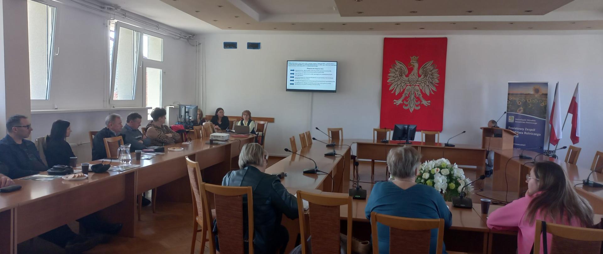 Spotkanie w sali konferencyjnej z udziałem kilkunastu osób siedzących przy stołach i oglądających prezentację wyświetlaną na ekranie. Na ścianie widoczne jest godło Polski, obok stoją flagi państwowe.