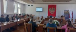 Spotkanie w sali konferencyjnej z udziałem kilkunastu osób siedzących przy stołach i oglądających prezentację wyświetlaną na ekranie. Na ścianie widoczne jest godło Polski, obok stoją flagi państwowe.