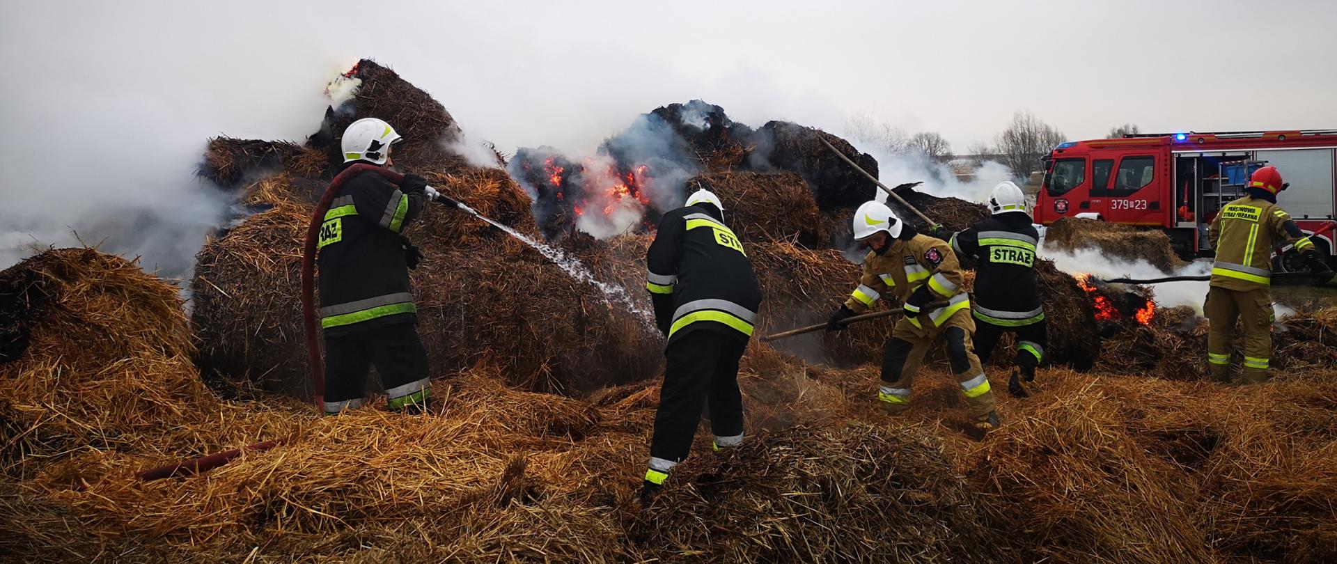 Zdjęcie przedstawia pożar słomy w balotach oraz strażaków którzy gaszą pożar przy użyciu wody oraz rozgarniają spaloną słomę. 