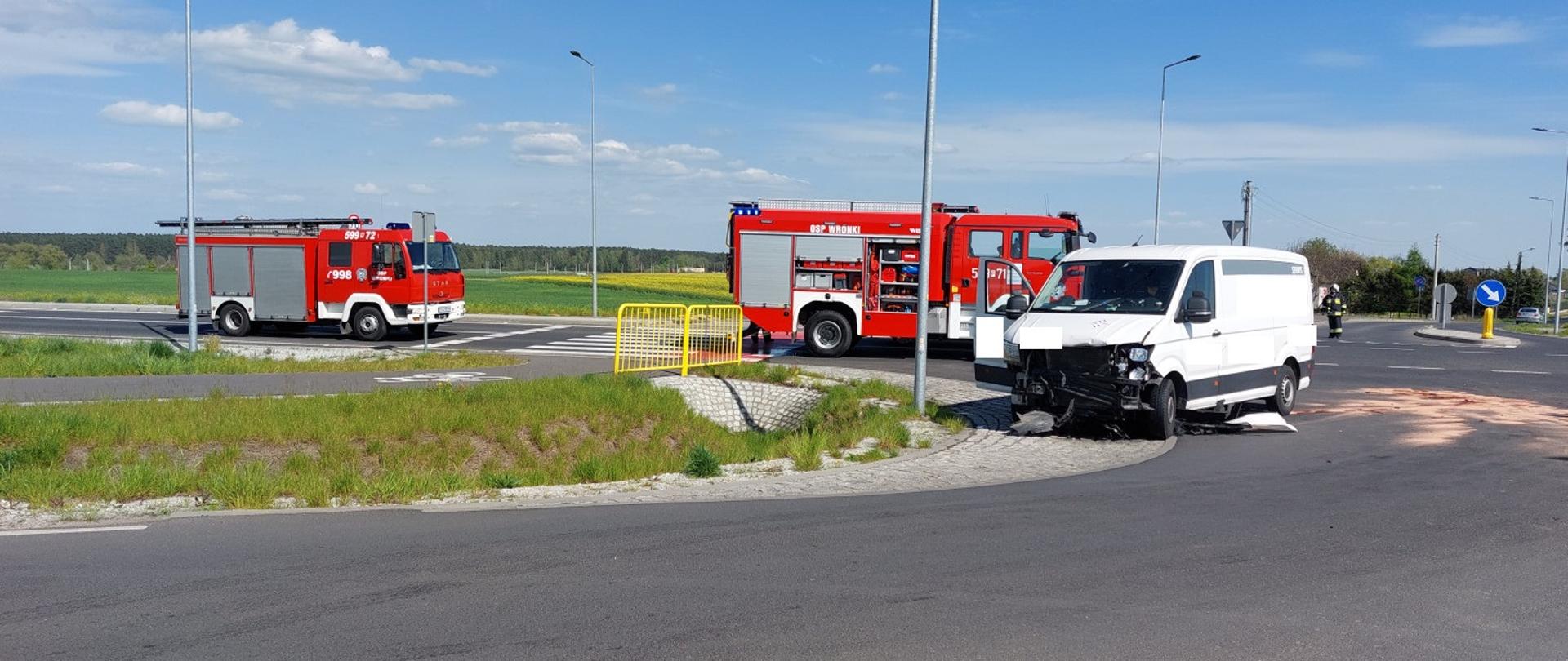 Widać uszkodzony biały samochód na skrzyżowaniu, za nim dwa samochody strażackie