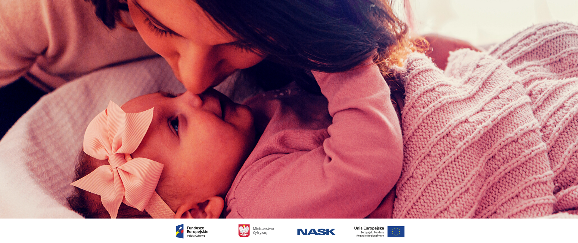 Matka pochylająca się nad córką leżącą w koszyku. Dziewczynka ma na głowie kokardkę. Zdjęcie z przewagą koloru różowego.