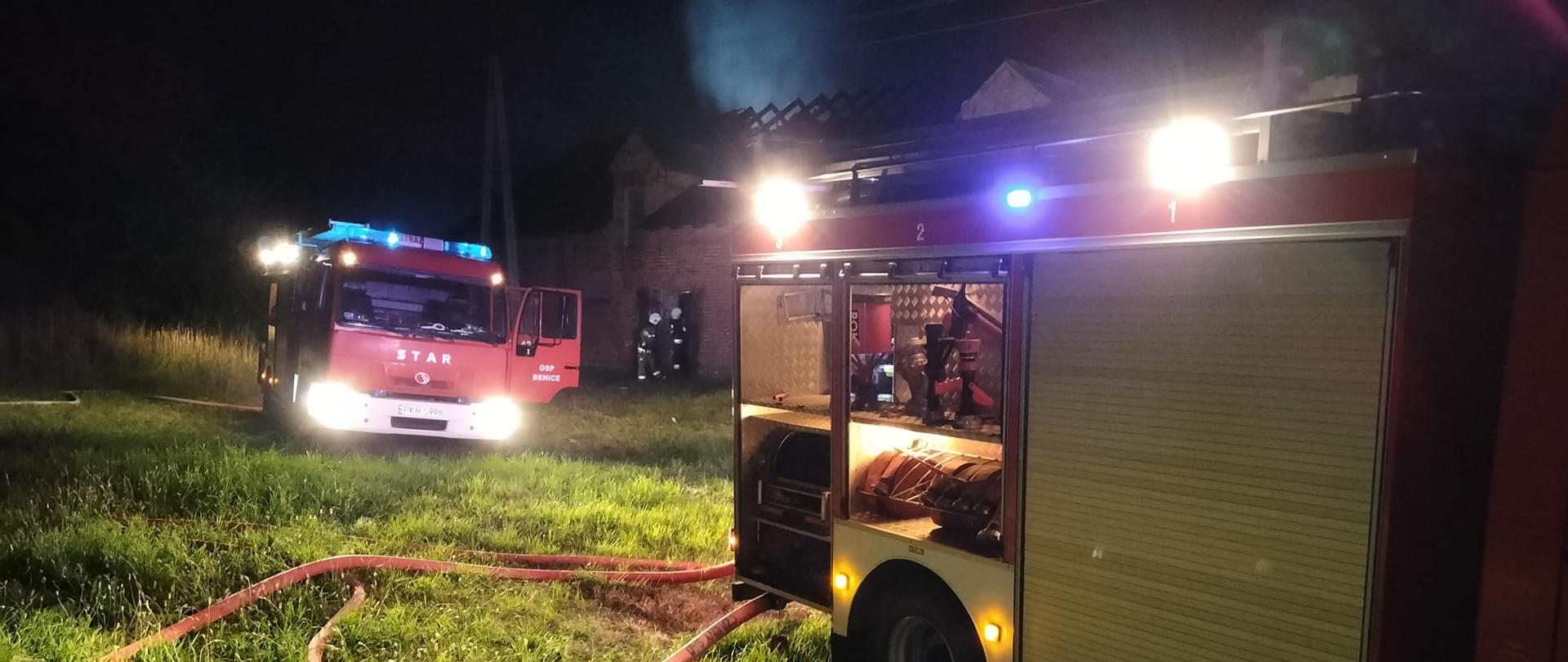 W pobliżu stodoły stoją samochody gaśnicze z włączonym oświetleniem zewnętrznym. Trwa akcja gaśnicza. Do samochodów podłączono węże pożarnicze. Jest noc. W tle widoczna stara stodoła ze spalonym dachem, nad którym unosi się dym.