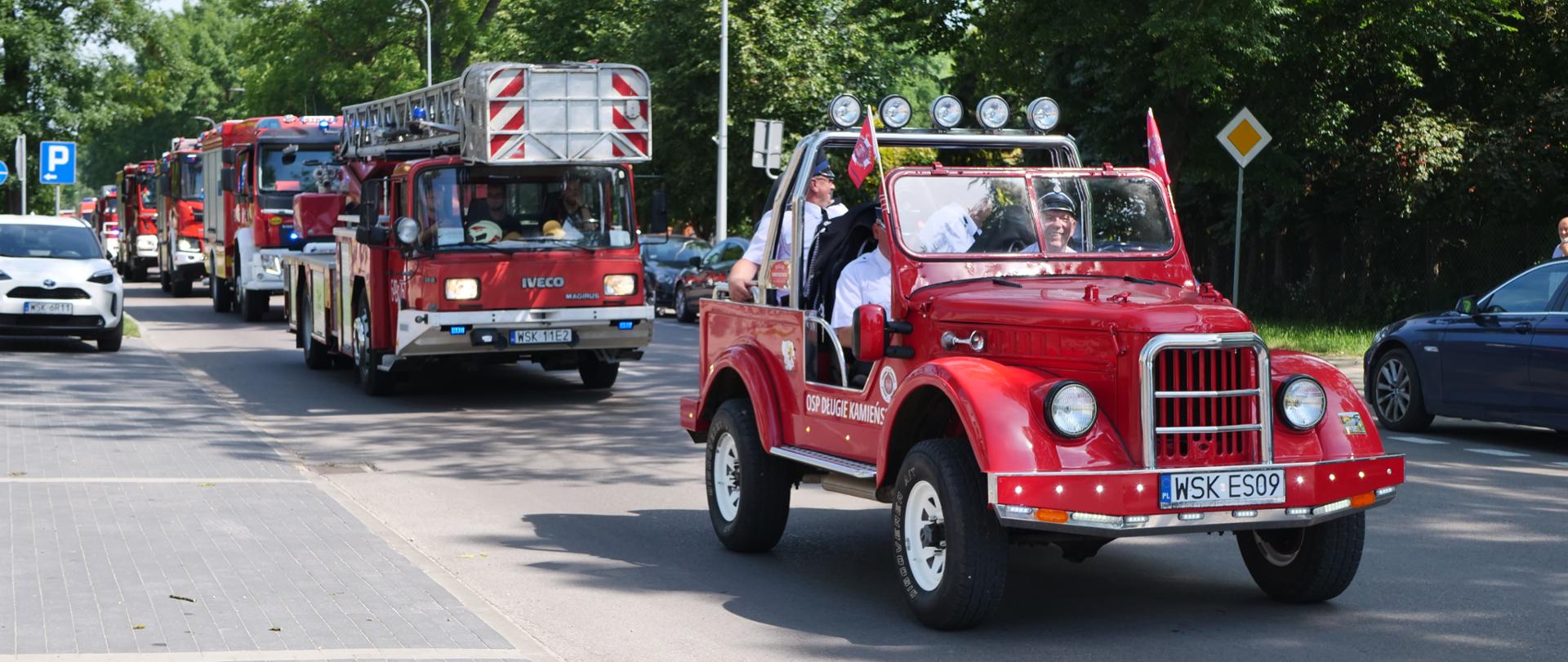 Zdjęcie przedstawia uroczystą paradę pojazdów pożarniczych poruszających się kolumną przez miasto. Na pierwszym planie jedzie czerwony, zabytkowy samochód gaśniczy bez dachu (typu retro/cabrio) z oznaczeniem „OSP DŁUGIE KAMIEŃSKIE”, w którym siedzą uśmiechnięci druhowie w białych koszulach. Za nim podąża nowoczesny wóz drabiniasty marki Iveco Magirus oraz inne współczesne samochody ratowniczo-gaśnicze na sygnale.