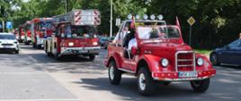 Zdjęcie przedstawia uroczystą paradę pojazdów pożarniczych poruszających się kolumną przez miasto. Na pierwszym planie jedzie czerwony, zabytkowy samochód gaśniczy bez dachu (typu retro/cabrio) z oznaczeniem „OSP DŁUGIE KAMIEŃSKIE”, w którym siedzą uśmiechnięci druhowie w białych koszulach. Za nim podąża nowoczesny wóz drabiniasty marki Iveco Magirus oraz inne współczesne samochody ratowniczo-gaśnicze na sygnale.