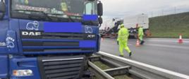Widać ciągnik siodłowy koloru niebieskiego, który uderzył przodem w bariery, po prawej widać dwóch pracowników obsługi autostrady