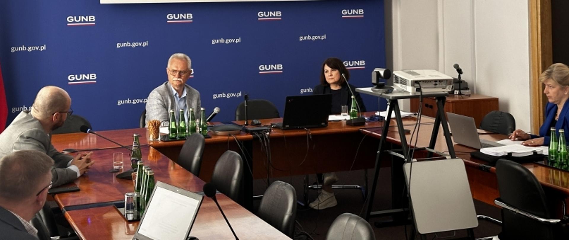 Pięć osób przy stole konferencyjnym podczas spotkania w GUNB, z banerem „gunb.gov.pl” i flagą Polski w tle.