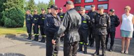 Komendant składa życzenia st. ogn. Arturowi Szymańskiemu. W tle stoją inni strażacy.