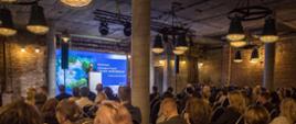 The frame shows a loft-style conference room with participants seated on chairs during a conference. On stage is a woman—Dorota Zawadzka-Stępniak, President of the NFEP&WM—with a large screen behind her.