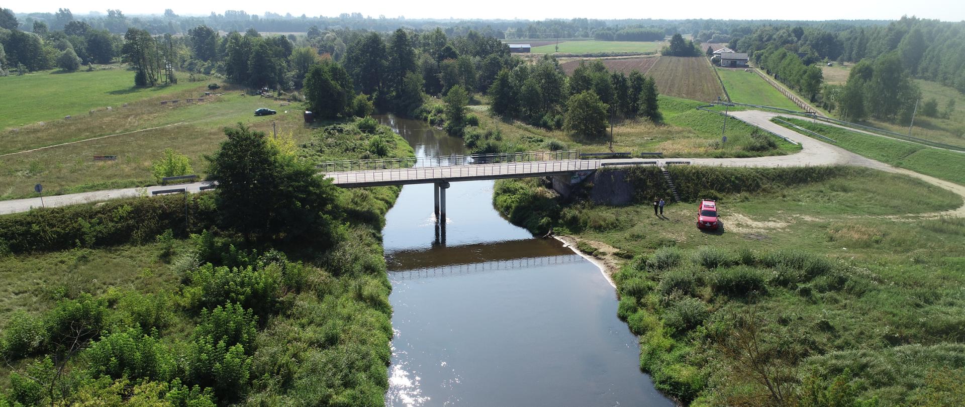 Zdjęcie wykonane z drona, widoczna rzeka, most obok którego stoi samochód specjalny strażacki. W oddali widoczne drzewa wzdłuż rzeki. 