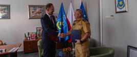 W pomieszczeniu z szarymi ścianami stoi prezydent miasta ubrany w granatowy garnitur oraz komendant w piaskowym mundurze. W rękach trzymają teczki z porozumieniem, ściskają sobie prawe dłonie. W tle flaga Polski.