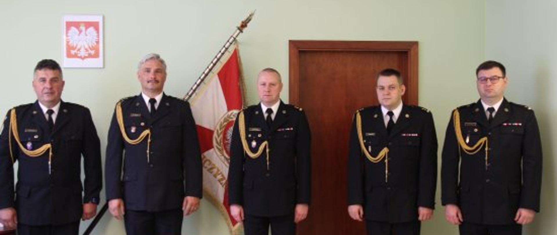 Zdjęcie przedstawia pięciu funkcjonariuszy w mundurach galowych na tle sztandaru oraz godła polskiego. od lewej brygadier Jacek Michalak komendant miejski Państwowej Straży Pożarnej w Poznaniu starszy kapitan Radosław Hurnik zastępca komendanta miejskiego Państwowej Straży Pożarnej w Poznaniu, Wielkopolski Komendant Wojewódzki Państwowej Straży Pożarnej starszy brygadier Dariusz Matczak, zastępca Wielkopolskiego komendanta wojewódzkiego brygadier Jarosław Zamelczyk oraz drugi zastępca Wielkopolskiego komendanta wojewódzkiego Robert Natunewicz