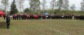 Na fotografii na płycie stadionu stoją w szeregu członkowie drużyn jednostek Ochotniczych Straży Pożarnej, które biorą udział w zawodach. Są ustawieni w grupach podzielonych na poszczególne gminy. Pierwszy zawodnik w grupie trzyma tablicę z nazwą gminy jaką zespoły reprezentują. Zawodnicy ubrani w umundurowanie polowe oraz dresy sportowe wymagane w zawodach. Na głowach zawodnicy mają ubrane hełmy strażackie. Zdjęcie wykonane podczas zbiórki przed zawodami. 