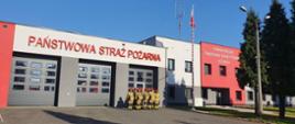 Strażacy w mundurach bojowych stoją w dwuszeregu na tle szarej ściany komendy. Po ich lewej stronie powiewa wciągnięta na maszt flaga państwowa.