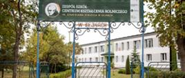 Brama wejściowa, nad bramą baner z wizerunkiem patrona oraz z nazwą szkoły, w tle główny budynek szkoły