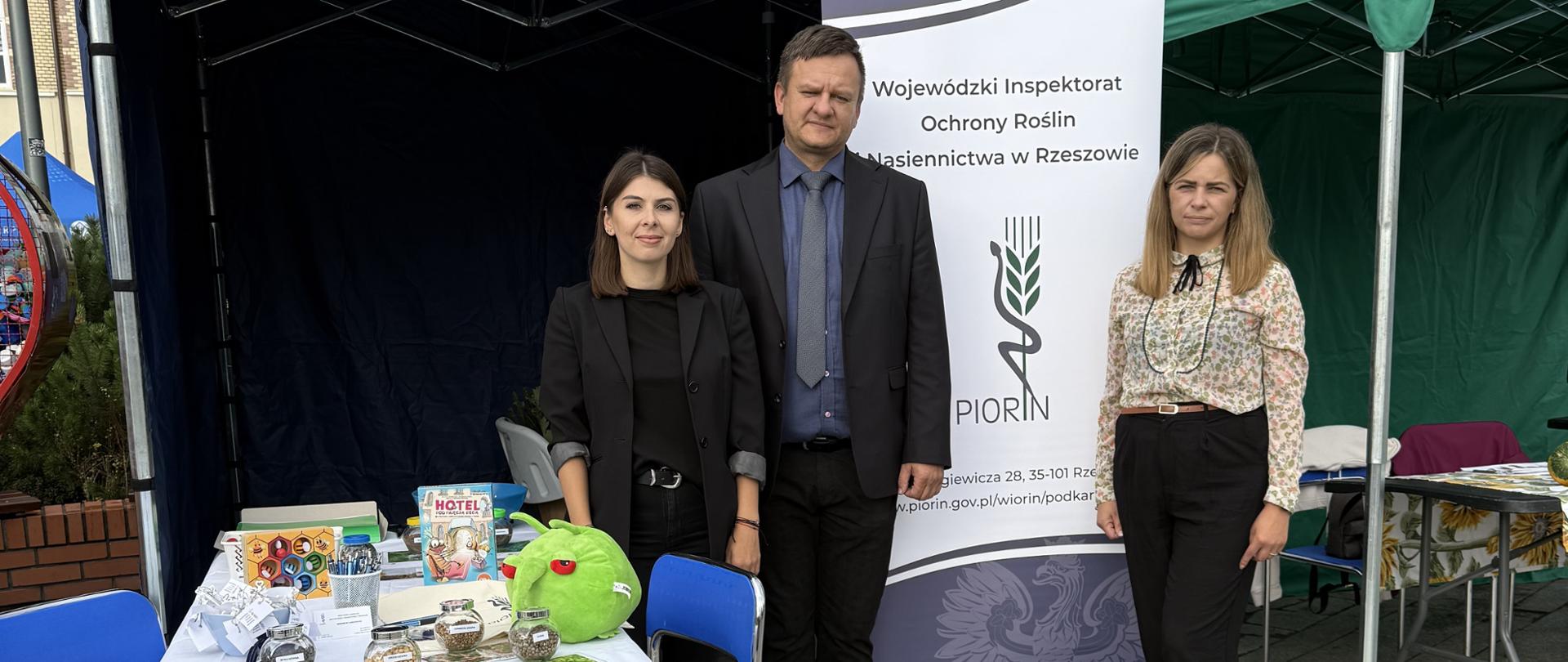 Trzy osoby -mężczyzna i dwie kobiety. Ubrani są elegancko. Stoją przed biało-granatowym banerem z logiem instytucji. Po lewej stronie jest stolik z różnymi materiałami.