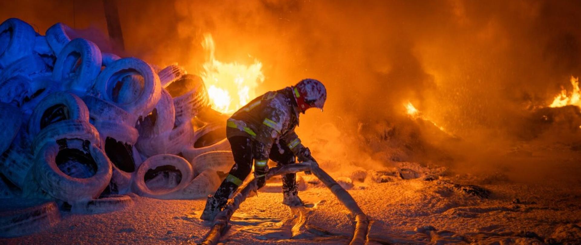 Zdjęcie z miejsca prowadzonych działań gaśniczych składowiska opon - widać na pierwszym planie strażaka, który przemieszcza wąż, zatopiony w warstwie piany gaśniczej. W tle łuna pożaru.