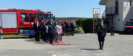 Zdjęcie przedstawia teren Jednostki Ratowniczo-Gaśniczej oraz dowódcę uroczystości ubranego w mundur galowy koloru czarnego ze sznurem galowym oraz czapką rogatywką na głowie, podczas obchodów Dnia Strażaka w Radziejowie. Na drugim planie przedstawiciele samorządów z terenu powiatu radziejowskiego oraz zaproszeni goście, zmiana służbowa strażaków oraz wozy bojowe straży pożarnej