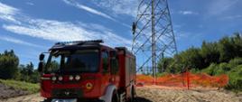 Na zdjęciu widać na pierwszym planie samochód pożarniczy, w tle widać słup elektroenergetyczny, na który wchodzi strażak celem dostania się do osoby poszkodowanej