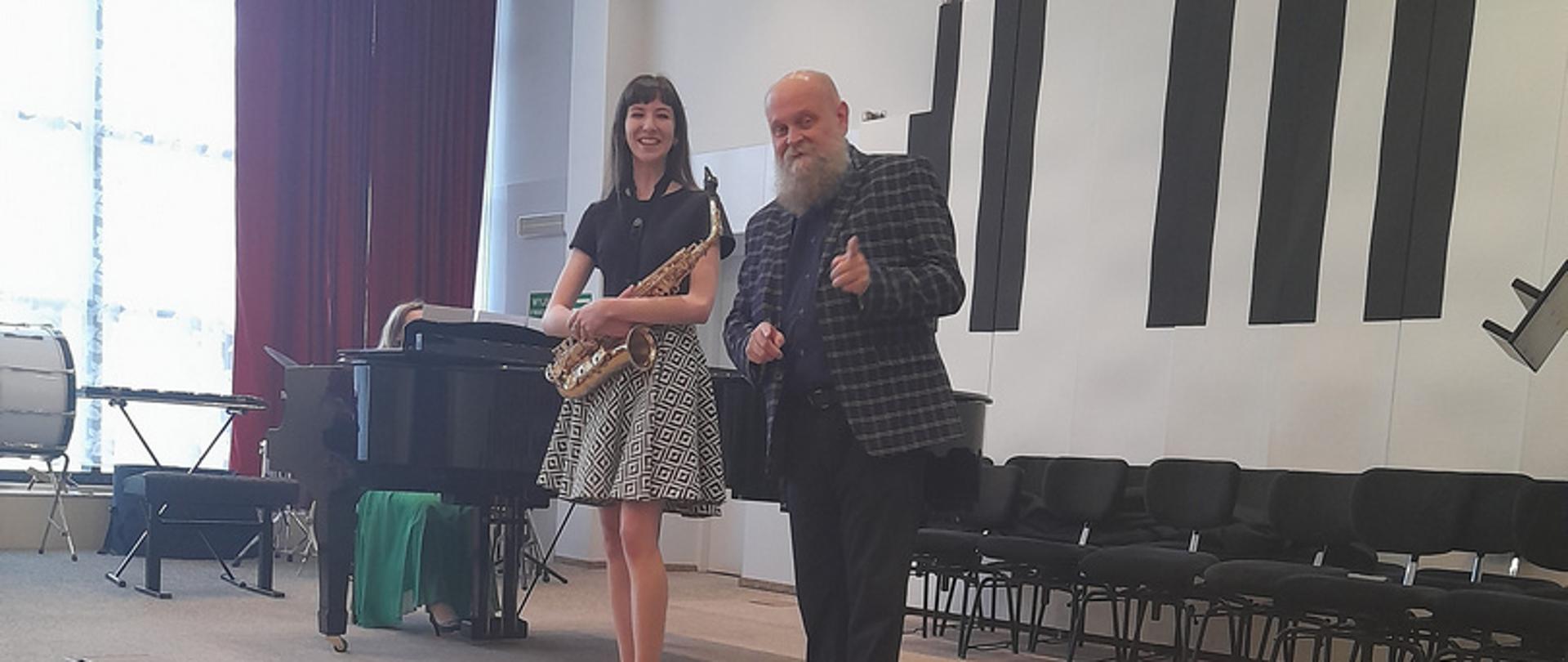 Na estradzie auli PSM stoi mężczyzna i nastolatka, która trzyma w rękach przed sobą saksofon.