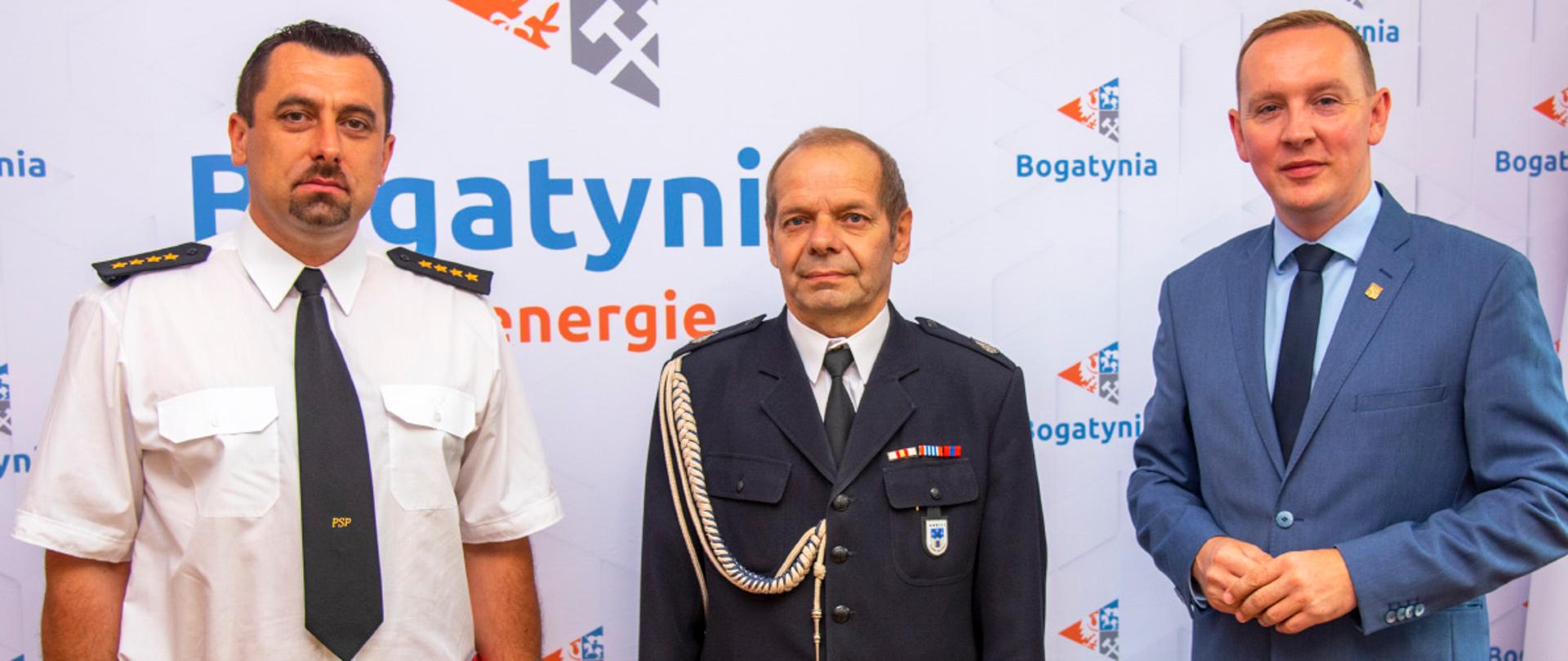 Na tle białej ściany na którzej znajdują się grafiki-loga Gminy Bogatynia oraz napsiy róznej wielkości w kolorze niebieskim "Bogatynia" stoją od lewej funkcjonariusz PSP ubrany w biała koszulę, czarny krawat ze złotym napisem "PSP" oraz czarne spodnie z brązowym paskiem. Na ramionach do koszuli przypięte pagony w kolorze czarnym z 4 złotymi gwiazdkami. W lewej dłoni trzyma teczkę papierową w kolorze czerwonym. Po jego lewej stronie stoi druh OSP ubrany w granatowy mundur galowy, srebrny sznur galowy, biała koszulę oraz czarny krawat. Na lewej kieszeni przypięty jakiś znaczek, nada kieszenią szereg baretek. Od prawej strony stoi mężczyzna ubrany w błękitny garnitur, jasno niebieską koszulę oraz czarny krawat