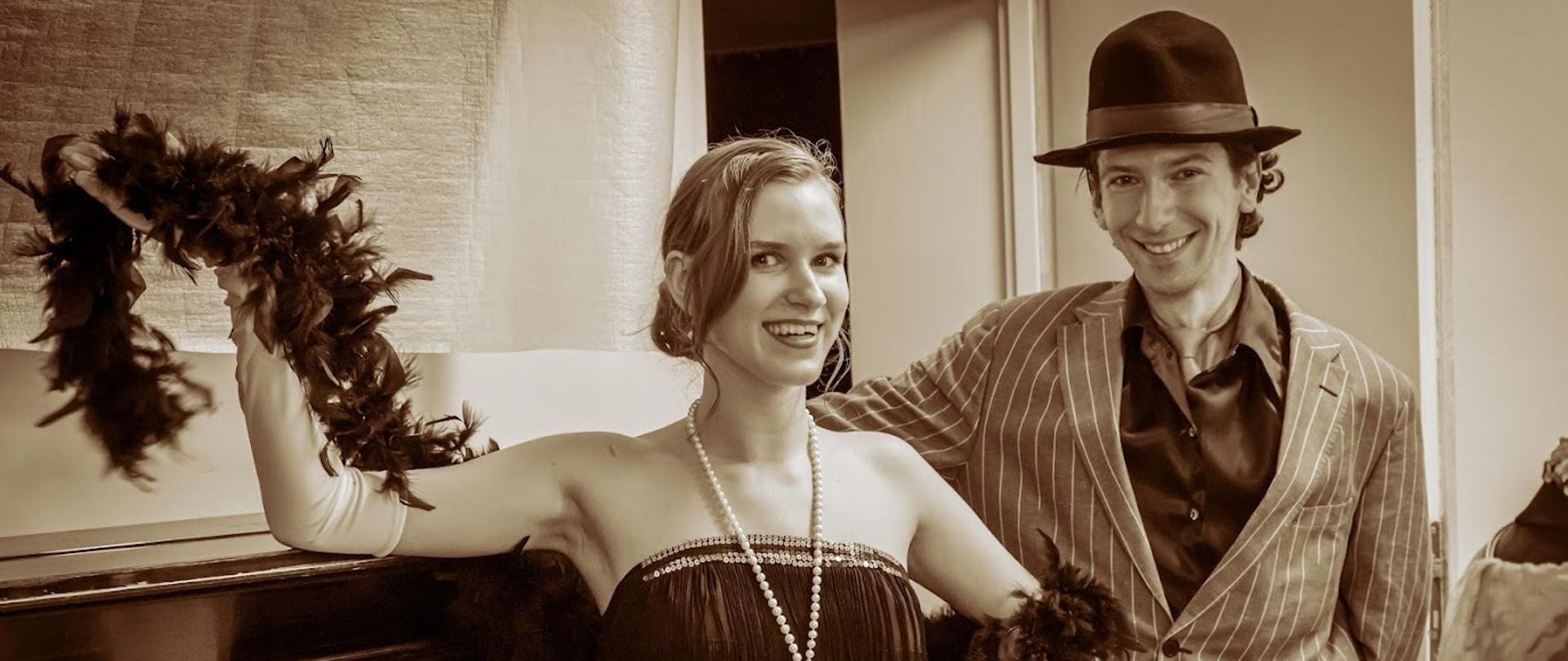 Zdjęcie w kolorze sepii. Na zdjęciu jest słuchaczka i słuchacz. słuchaczka ubrana jest w sukienkę, korale na szyi i boa z plecach. słuchacz ma marynarkę w paski i kapelusz na głowie. 