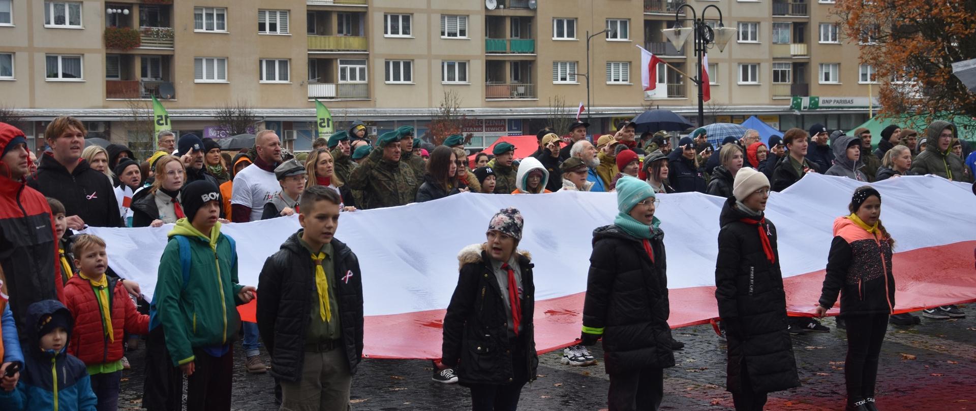 Zdjęcie przedstawia funkcjonariuszy Komendy Powiatowej Państwowej Straży Pożarnej w Gołdapi, którzy o godz. 12:00 na Placu Zwycięstwa w Gołdapi (nieopodal Pomnika Niepodległości) wspólnie z mieszkańcami miasta oraz przedstawicielami władz samorządowych i innych służb zaśpiewali hymn państwowy w ramach akcji „Niepodległa do Hymnu”. Uczestnicy uroczystości trzymają flagę Rzeczypospolitej Polskiej