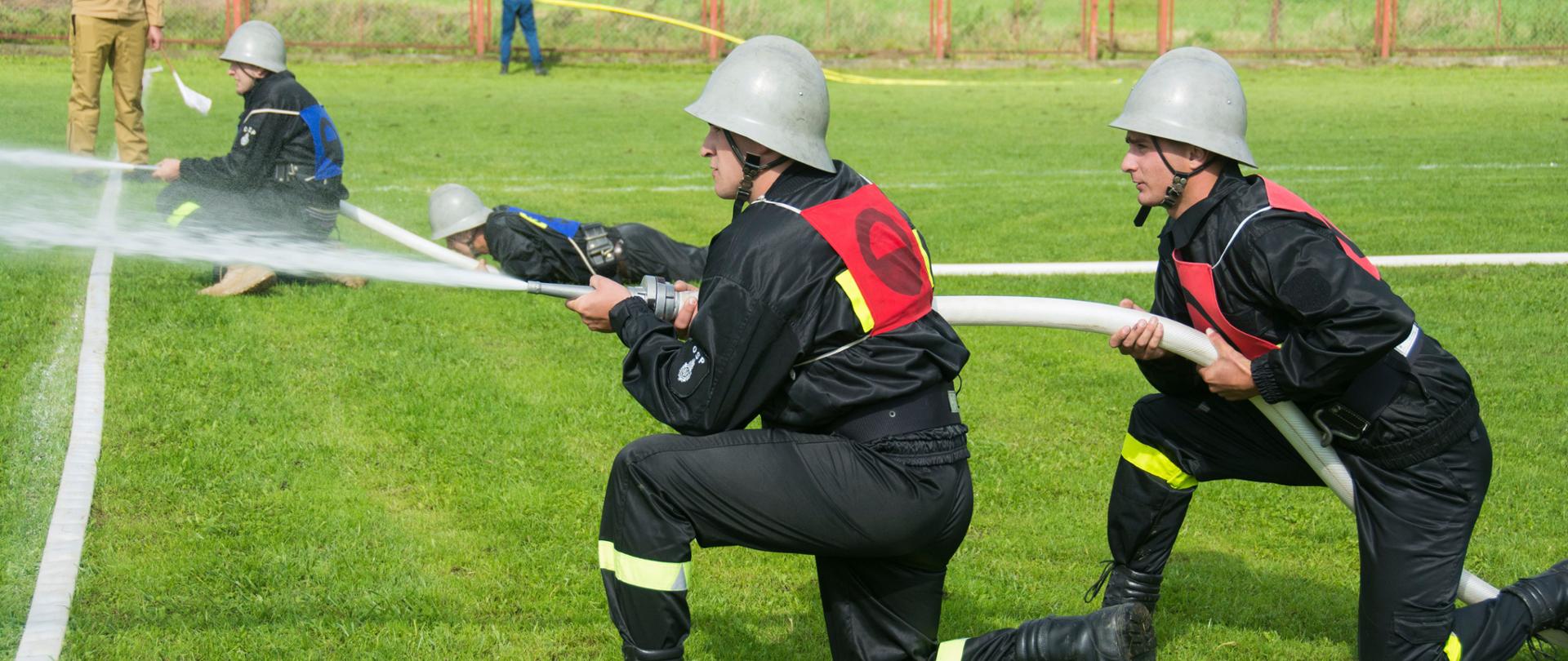 Kolorowa fotografia wykonana w pochmurny dzień na zewnątrz. Przedstawia strażaków OSP ubranych w hełmy i ubrania koszarowe podczas zawodów sportowo pożarniczych w czasie ćwiczenia bojowego. Na drugim planie sędzia zawodów w jasnym ubraniu służbowym.