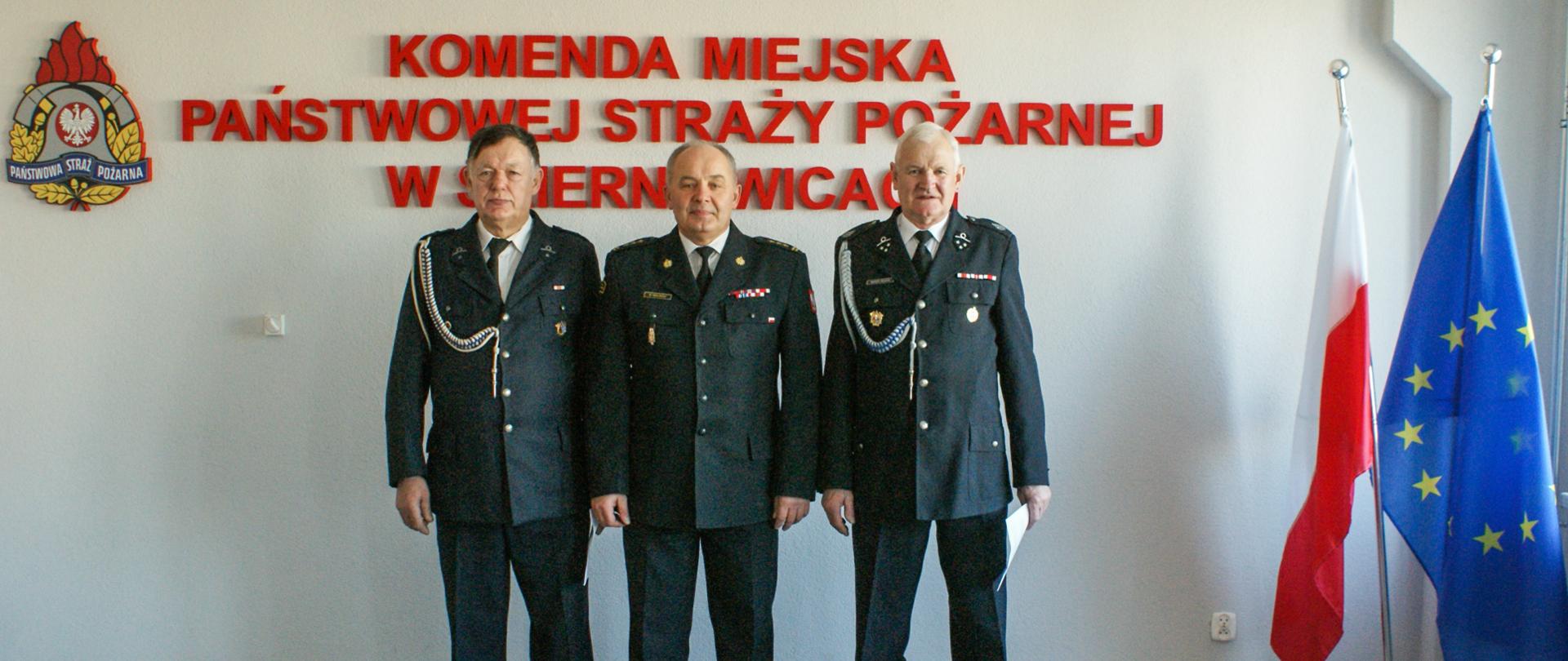 Zdjęcie przedstawia trzech strażaków w mundurach wyjściowych koloru czarnego na tle czerwonego napisu Komenda Miejska Państwowej Straży Pożarnej w Skierniewicach, loga psp, flag polski i unii europejskiej.
