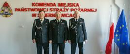 Zdjęcie przedstawia trzech strażaków w mundurach wyjściowych koloru czarnego na tle czerwonego napisu Komenda Miejska Państwowej Straży Pożarnej w Skierniewicach, loga psp, flag polski i unii europejskiej.