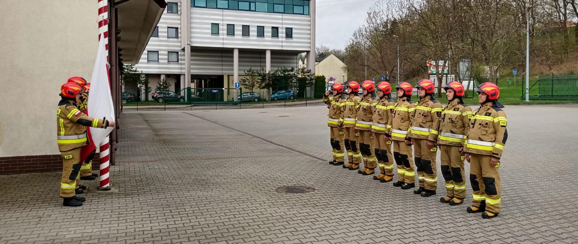 Na zdjęciu widoczny na placu wewnętrznym przed Komendą PSP w Gorlicach poczet flagowy przy maszcie w ubraniach specjalnych w kolorze piaskowym i czerwonych hełmach podczas podnoszenia flagi w zawiązku z uroczystością Dnia Flagi Rzeczypospolitej Polskiej. Przed pocztem ustawiona zmiana służbowa w szeregu w pozycji na baczność również w ubraniach specjalnych koloru pisakowego i czerwonych hełmach. 