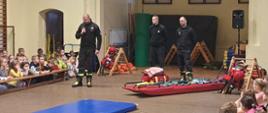 Trzech stojących wewnątrz hali sportowej umundurowanych strażaków. Jeden z nich mówi do mikrofonu. Przed strażakami rozłożony sprzęt ratowniczy. Wewnątrz hali na podłodze siedzą dzieci.
