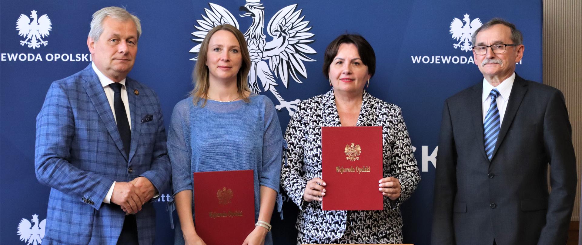 Dwie kobiety i dwóch mężczyzn na niebieskim tle. Kobiety trzymają przed sobą czerwone teczki.