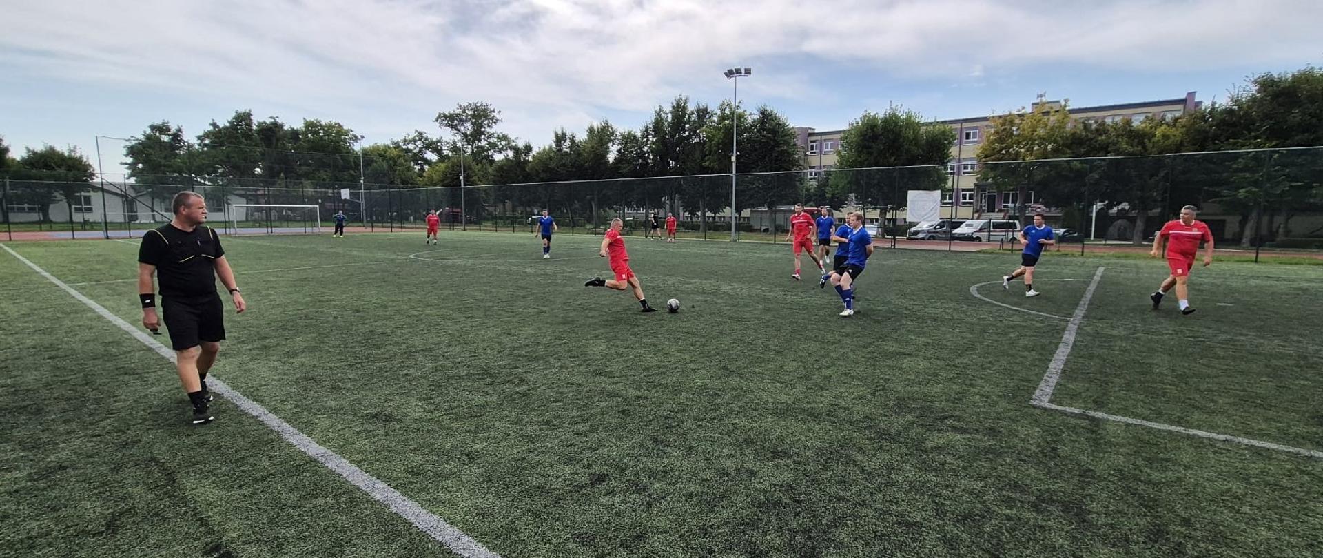 Na zdjęciu widać mecz piłki nożnej rozgrywany na boisku ze sztuczną murawą. Drużyna w czerwonych strojach gra przeciwko drużynie w niebieskich koszulkach. W centrum akcji zawodnik w czerwonym stroju kopie piłkę, a kilku zawodników w niebieskich koszulkach stara się go zatrzymać. Z lewej strony kadru znajduje się sędzia w czarnym stroju, obserwujący przebieg gry. W tle widać bramkę, ogrodzenie boiska, drzewa oraz budynki mieszkalne.