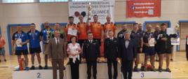 Zdjęcie przedstawia trzy drużyny strażaków z województwa podkarpackiego, śląskiego i opolskiego, stojących na podium w hali sportowej podczas XXXVII Mistrzostw Polski Strażaków Państwowej Straży Pożarnej w Tenisie Stołowym. Strażacy, którzy zajęli odpowiednio 1, 2 i 3 miejsce trzymają w rękach dyplomy i puchary. 