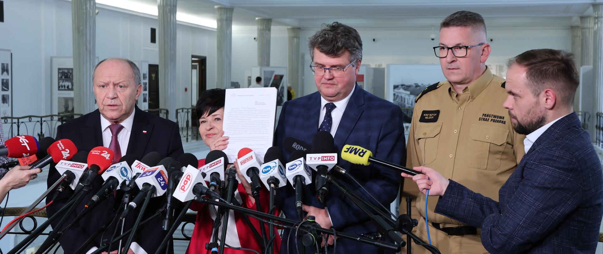 Przedstawiciele parlamentu (2 osoby) wiceminister MSWiA oraz z-ca KG PSP stoją przy mikrofonach podczas konferencji prasowej