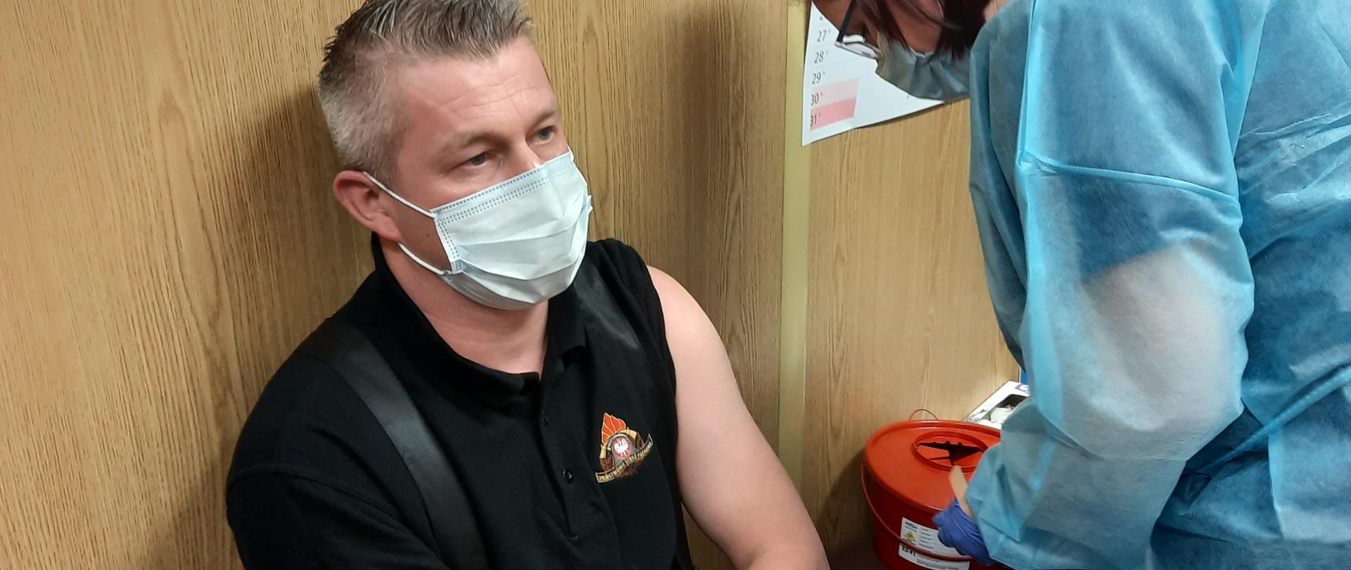 Zdjęcie przedstawia mężczyznę w średnim wieku w mundurze funkcjonariusza Państwowej Straży Pożarnej siedzącego na ławeczce z podwiniętym lewym rękawem koszulki, oczekującego na szczepienie. Na zdjęciu znajduje się też pielęgniarka w niebieskim fartuchu przygotowująca zastrzyk.