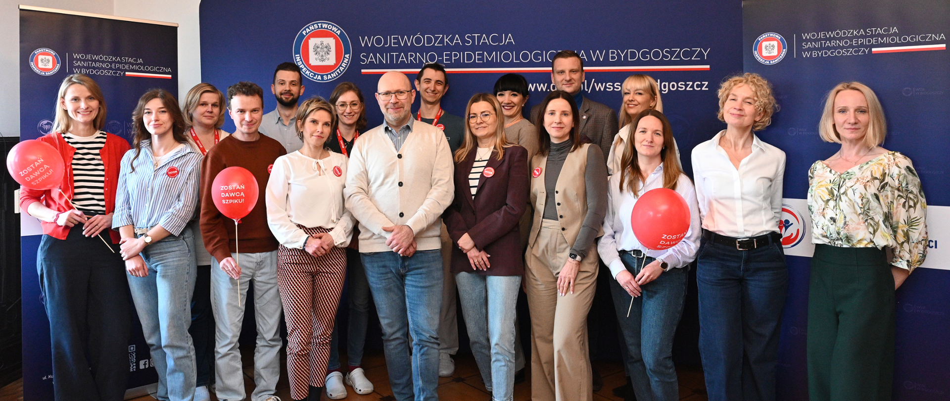 Grupa kilkunastu osób pozuje do wspólnego zdjęcia we wnętrzu urzędu na tle granatowych banerów Wojewódzkiej Stacji Sanitarno-Epidemiologicznej w Bydgoszczy. Część uczestników trzyma czerwone balony z napisem „Zostań dawcą szpiku”, co sugeruje akcję społeczną lub kampanię informacyjną. Uczestnicy są ubrani półformalnie i stoją w dwóch rzędach, patrząc w stronę aparatu.
