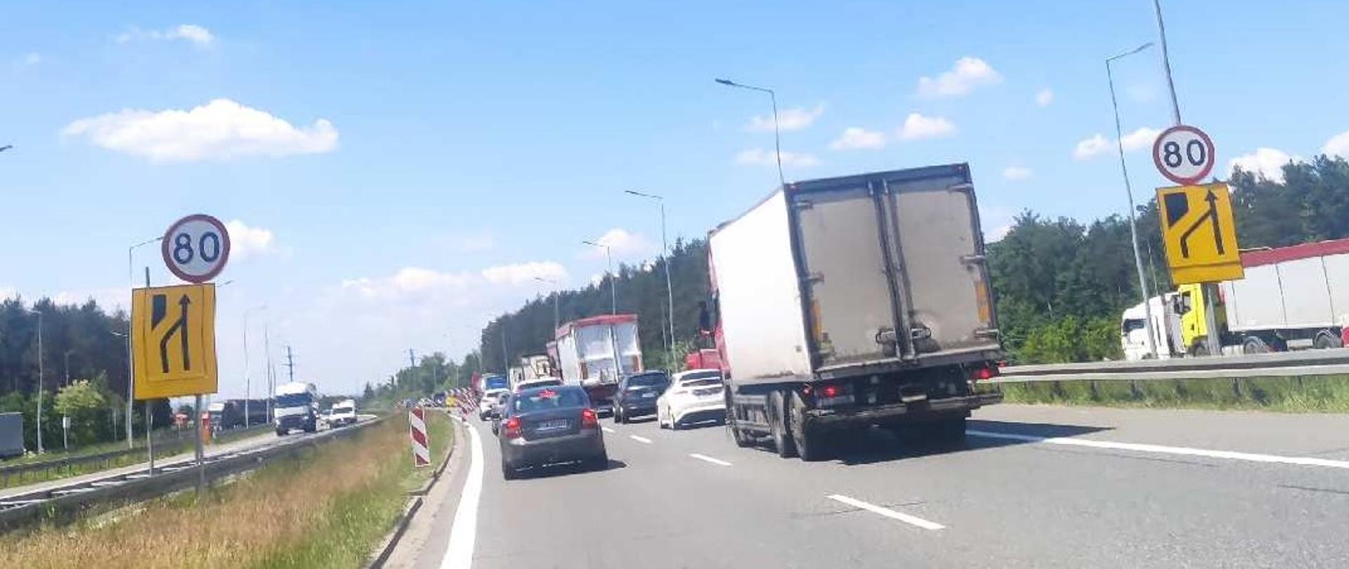 Zdjęcie przedstawia dwie drogi dwujezdniowe, trwale od siebie oddzielone pasem zieleni i barierą energo-chłonną. Widać ruch samochodów osobowych i ciężarowych w obu kierunkach. Zdjęcie zostało wykonane przez przednią szybę auta w słoneczny letni dzień. Po obydwu stronach jezdni widać znaki ograniczenia prędkości do 80 km a poniżej tych znaków, znaki informujące o zwężeniu pasa lewego. 