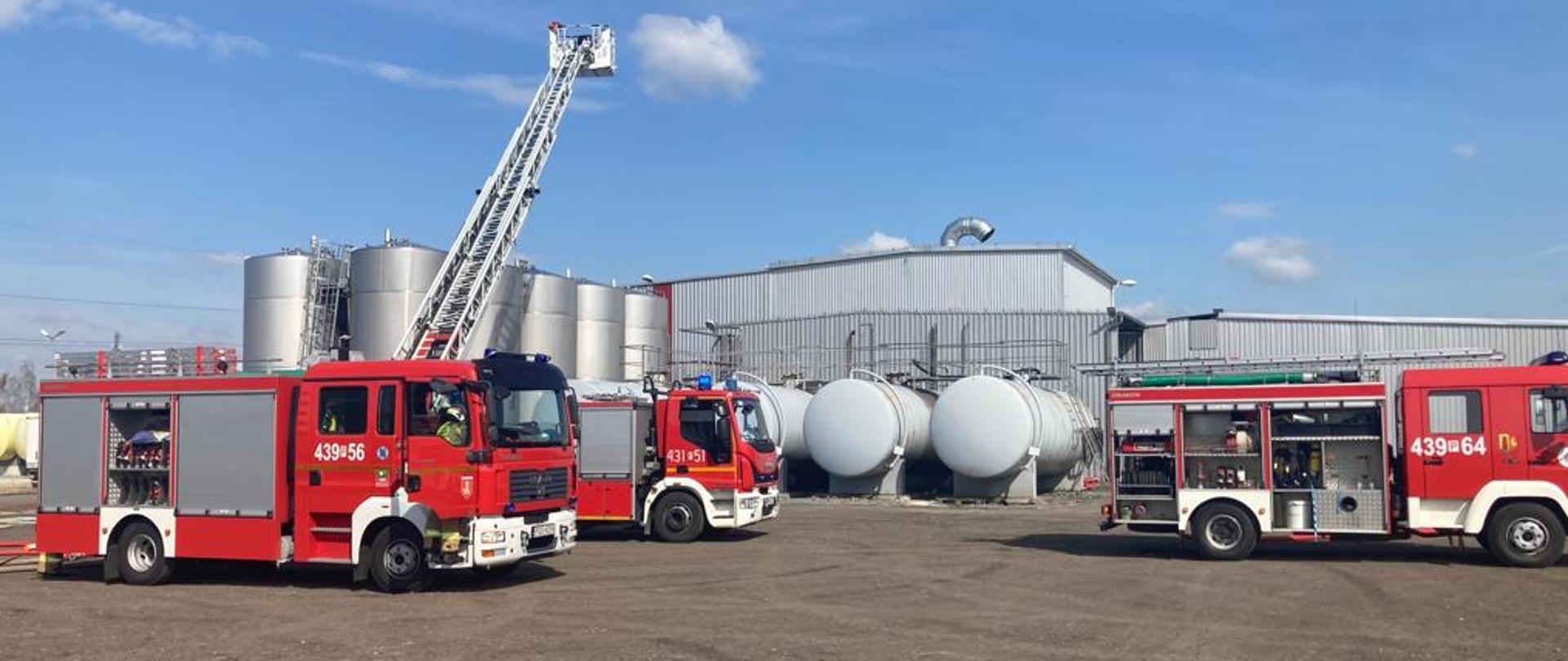 Zdjęcie przedstawia 3 wozy strażackie podczas ćwiczeń, w tym drabinę mechaniczną. Kosz drabiny wyciągnięty jest ponad zbiorniki technologiczne.