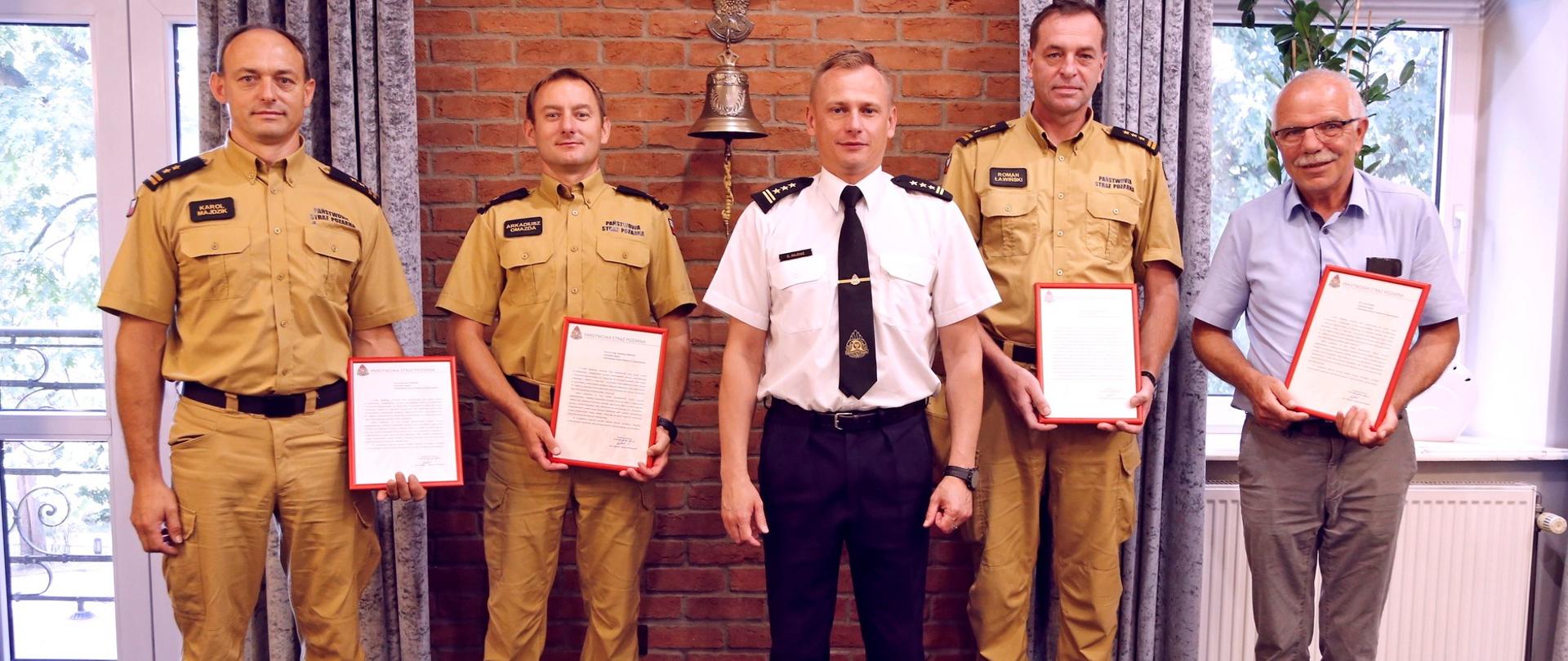 Grupowe zdjęcie Zastępcy Komendanta CS PSP (stojący w środku) po jego lewej stronie dwóch oficerów CS PSP trzymających w dłoni otrzymane podziękowanie, po jego prawej stronie oficer oraz pracownik fizyczny CS PSP prezentujący trzymane w dłoniach podziękowanie