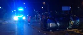 Zdjęcie przedstawia przewrócony na bok samochód osobowy - taksówka. Obok samochodu stoi strażak, za nim widać niebieską poświatę od sygnalizacji świetlnej samochodu straży pożarnej. Na jezdni widać porozrzucane pozostałości po wypadku.
