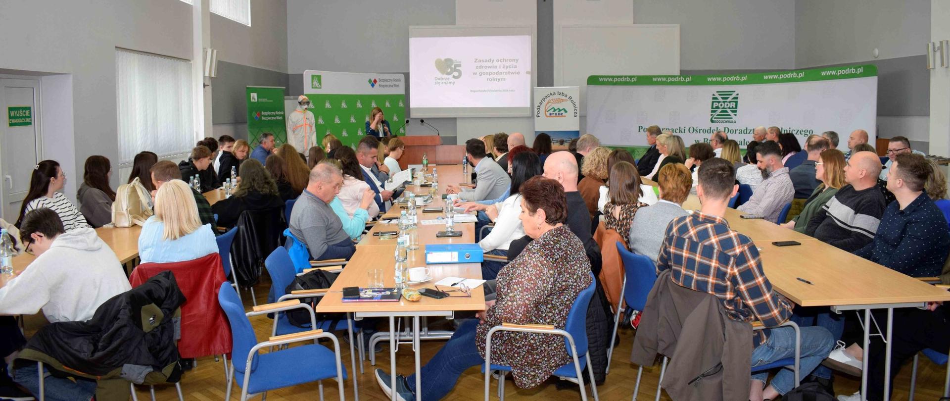 Duża, jasna sala konferencyjna wypełniona uczestnikami siedzącymi przy ustawionych w rzędach stołach. Na środku, przy mównicy, stoi osoba prowadząca prezentację, a obok niej manekin ubrany w strój ochronny, prawdopodobnie związany z tematyką rolniczą lub chemiczną. Na ekranie za prelegentem widoczny jest slajd z dużą liczbą „35” oraz tytułem dotyczącym zasad ochrony zdrowia i życia w gospodarstwie rolnym. Uczestnicy – kobiety i mężczyźni w różnym wieku – słuchają, czytają materiały lub notują. Na stołach znajdują się butelki wody, dokumenty i teczki. Ściany zdobią logotypy instytucji rolniczych, w tym Podkarpackiego Ośrodka Doradztwa Rolniczego.