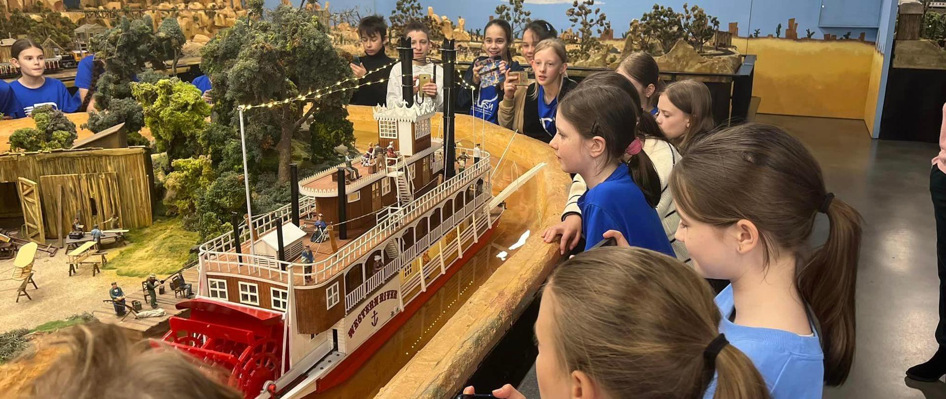 Grupa uczniów na tle zminiaturyzowanego świata ręcznie wykonanego zrobionego w studiu, na pierwszym planie statek w kolorze czerwono - biało - brązowym.