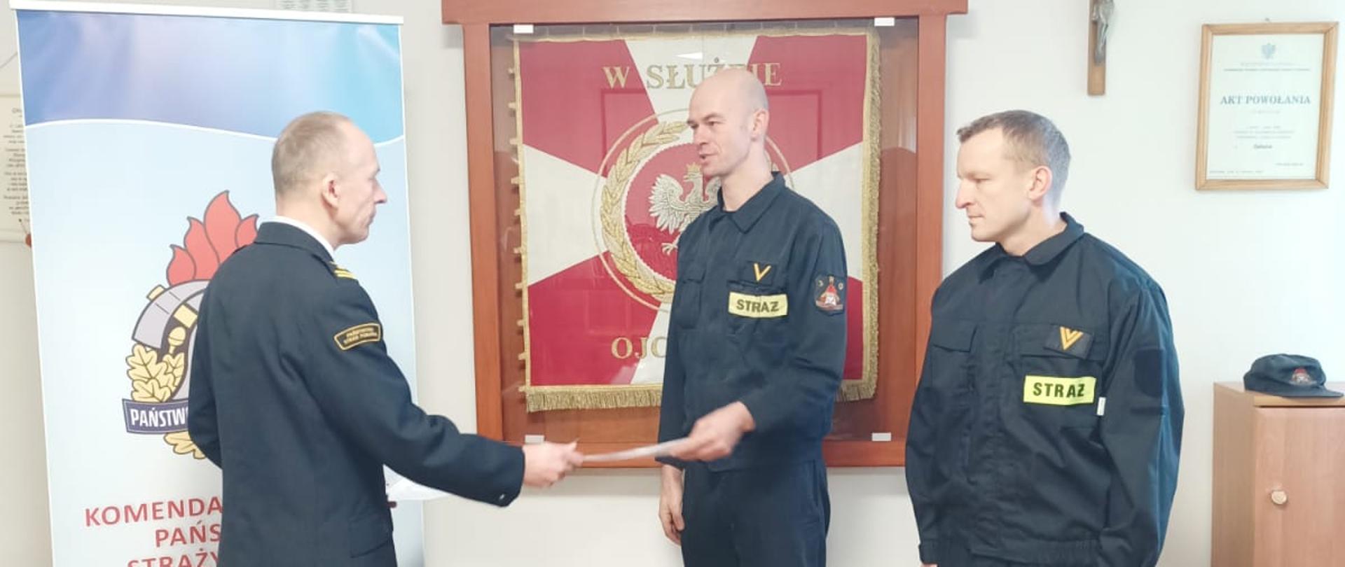 Zdjęcie przedstawia trzech funkcjonariuszy podczas wręczania awansów w świetlicy. Za nimi znajduje się sztandar KP PSP Świdwin, godło oraz krzyż.