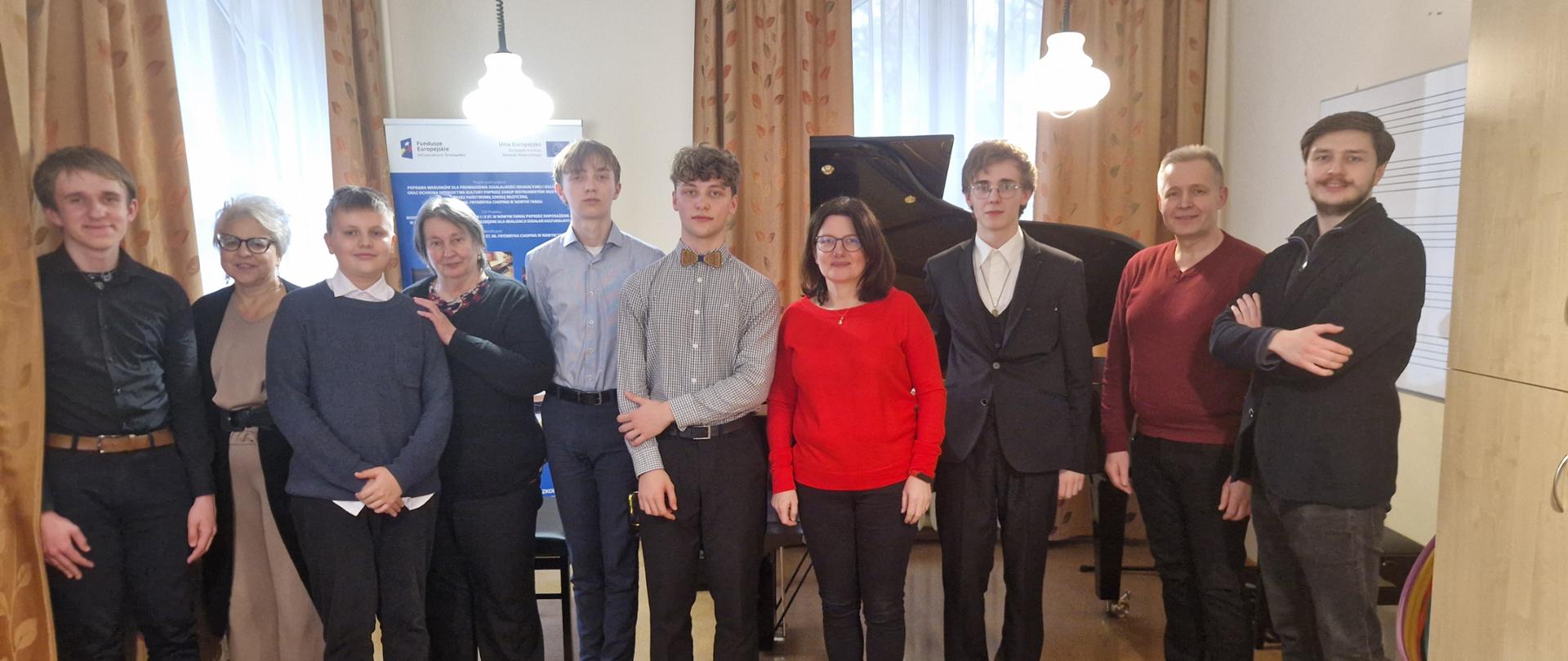 Zdjęcie grupowe. Na sali w jednym rzędzie stoją uczniowie oraz nauczyciele fortepianu. Przed nimi puste krzesła, za nimi dwa okna przysłonięte firanami oraz zasłonami, baner unijny, fortepian, Z prawej strony tablica z pięciolinią oraz dwudrzwiowa szafa. Z sufitu zwisają zapalone lampy.