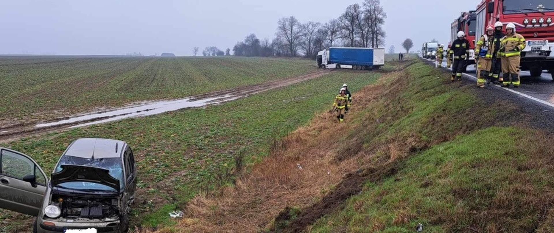 Po lewej stronie na polu stoją samochody biorące udział w zdarzeniu. Na drodze stoją pojazdy służb.