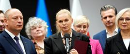 Minister Nowacka stoi przed mikrofonem na stojaku, w ręku trzyma fioletową teczkę, dookoła niej kilka osób.