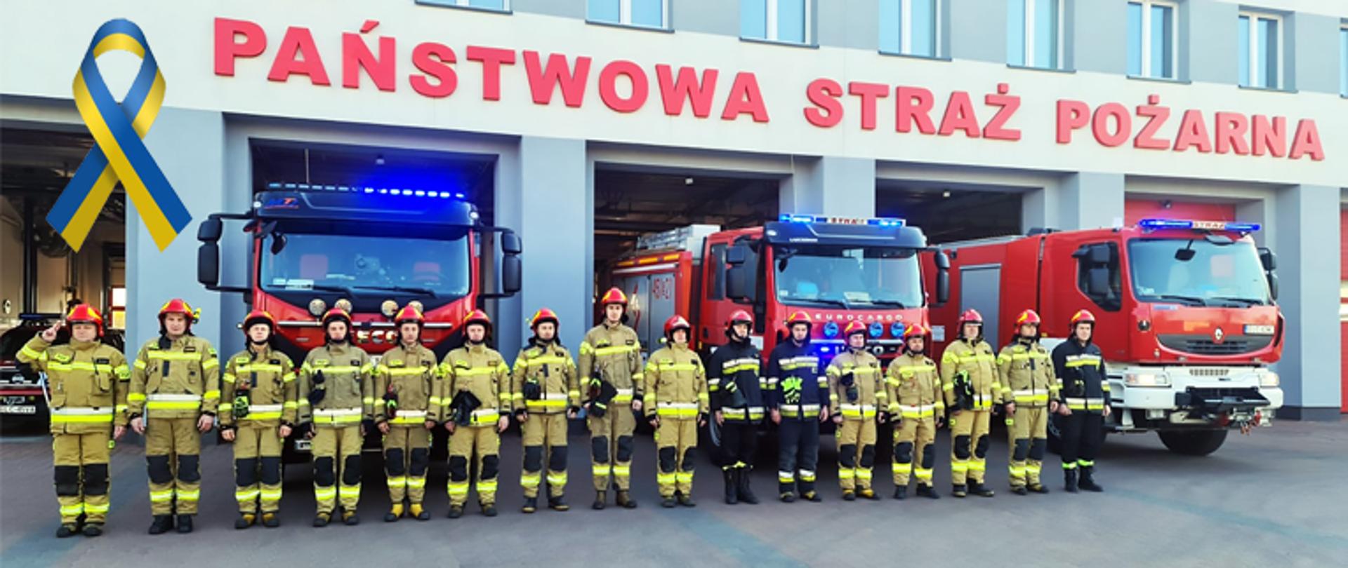Strażacy ubrani w ubranie bojowe typu moratex barwy jasnożółtej oraz w hełm barwy czerwonej stoją przed garażami, za ich plecami pojazdy gaśnicze barwy czerwonej z włączonymi światłami błyskowymi barwy niebieskiej, strażacy stoją w pozycji zasadniczej, strażak z lewej strony oddaje honor do hełmu, pojazdy wysunięte są przednią osią poza obrys bramy