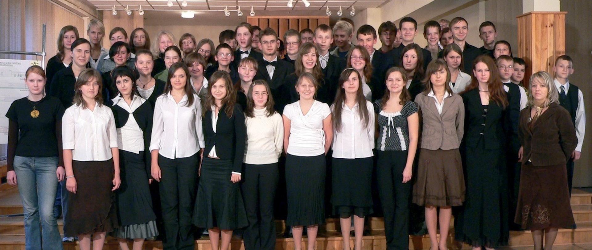 Młodzież szkolna stojąca w kilku rzędach. Z przodu dziewczęta w białych bluzkach i czarnych spódnicach. Z tyłu chłopcy w białych koszulach i czarnych marynarkach z muszkami. Z prawej strony stoi dyrygentka w brązowym kostiumie z apaszką.